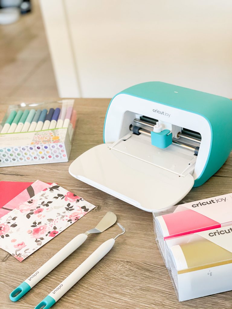 A Cricut Joy cutting machine is on a wooden table, surrounded by colorful pens, patterned paper, vinyl rolls, and crafting tools, ready for a creative project.