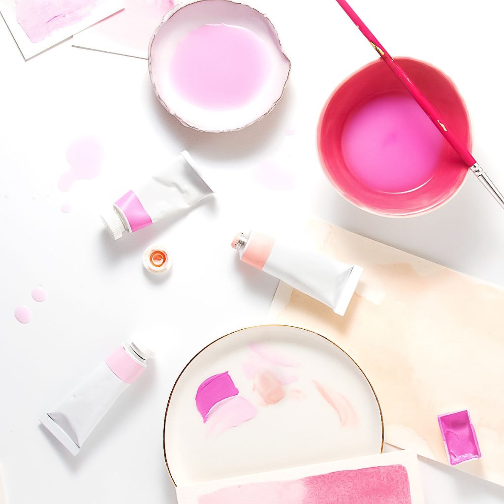 Art supplies on a white surface, including watercolor tubes, a paintbrush, bowls with pink water, paint swatches, watercolor paper, and paint stains in shades of pink and purple.