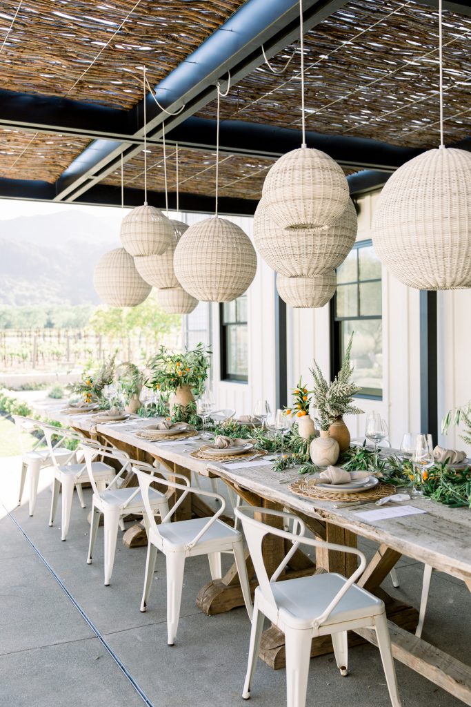 A rustic outdoor dining table set with greenery, neutral tableware, and earthy decor sits under a pergola. White paper lanterns hang overhead, and white chairs line both sides of the wooden table.