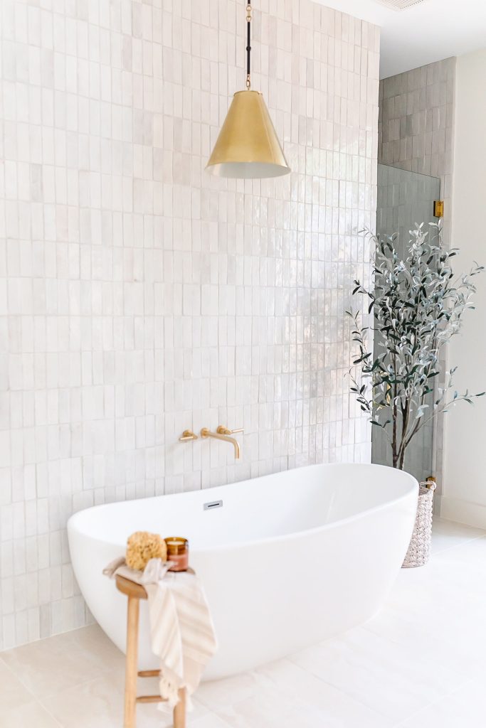 A modern bathroom with a white freestanding bathtub, gold fixtures, a wooden stool with a towel and bath accessories, a potted plant, and a gold pendant light against a tiled wall.