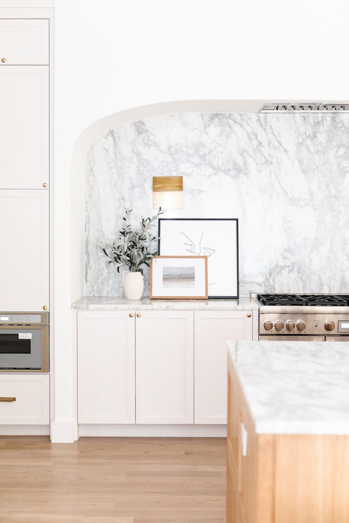 A bright, modern kitchen with white cabinets, marble countertops and backsplash, light wood floors, and minimal decor including framed art, a vase with greenery, and a gold wall sconce above the counter.