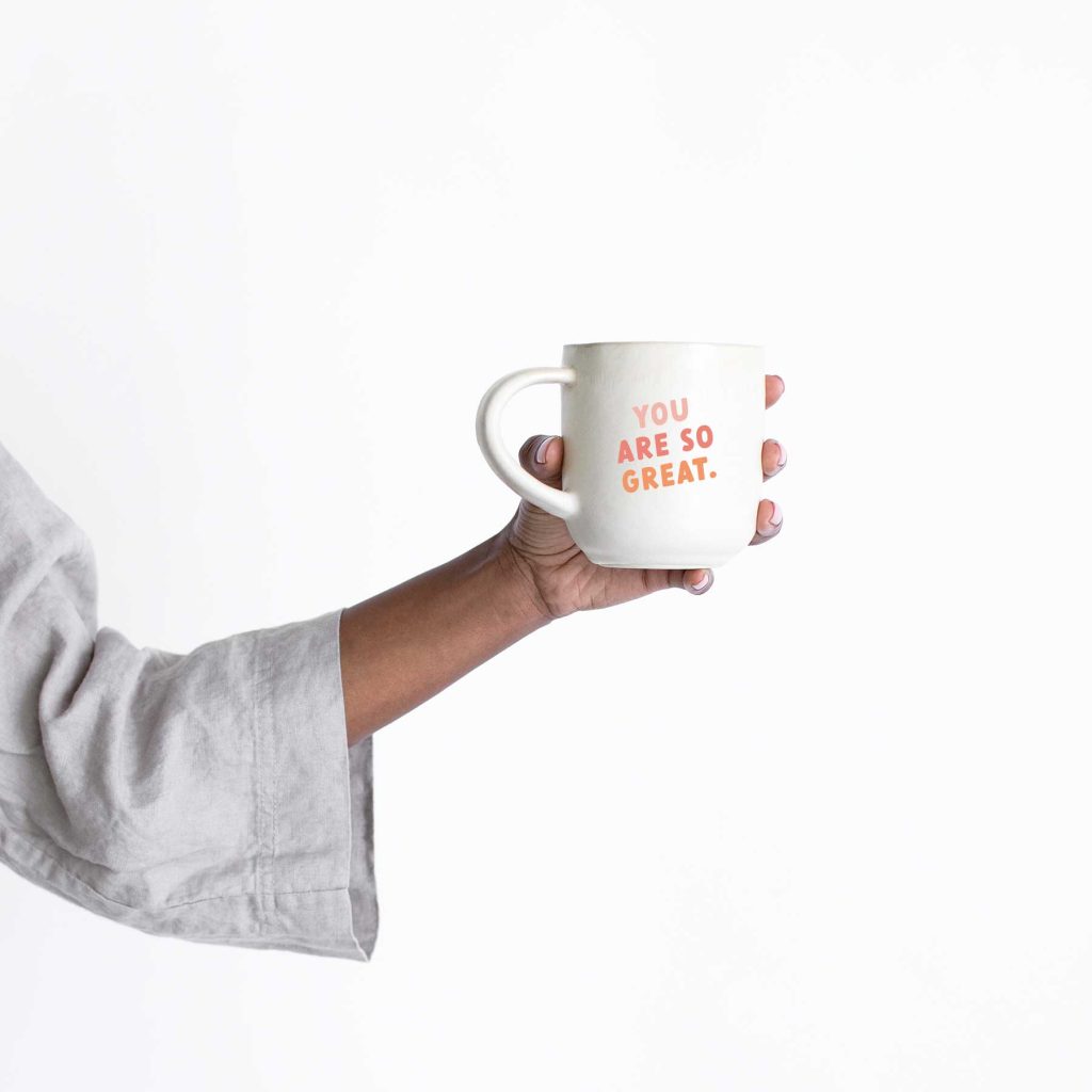 A person in a light gray sleeve holds a white mug with orange text that reads, YOU ARE SO GREAT, against a plain white background.