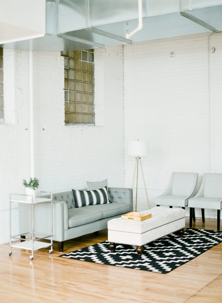 A modern, minimalist living room with a gray sofa, two gray chairs, a white ottoman, and a geometric black-and-white rug. A small rolling cart, standing lamp, and large windows with glass blocks complete the bright space.