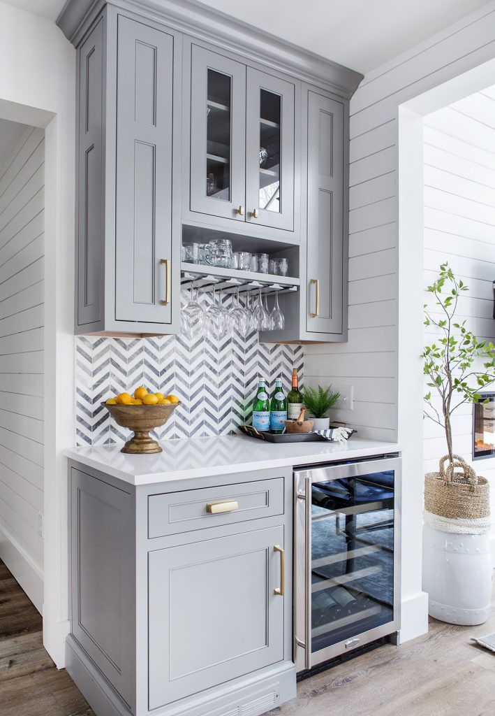 A small home bar with light gray cabinets, a white countertop, chevron-patterned backsplash, glassware, drinks, a bowl of lemons, and a wine fridge, set against white shiplap walls and a wood floor.