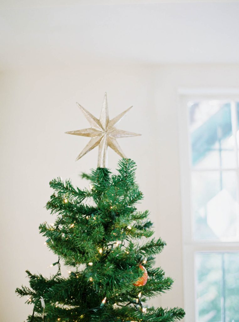 The top of a decorated Christmas tree with a gold star topper, lights, and ornaments, positioned in front of a bright window in a softly lit room.