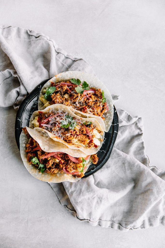 Three tacos filled with seasoned meat, onions, cheese, and fresh cilantro are arranged on a dark plate, set atop a light, textured cloth on a white surface.