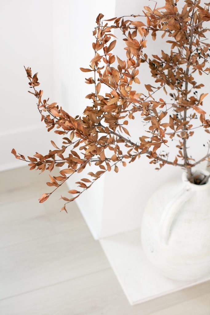 A white ceramic vase holds branches with dried brown leaves, placed on a light wooden floor against a white wall, creating a minimal and natural decor scene.