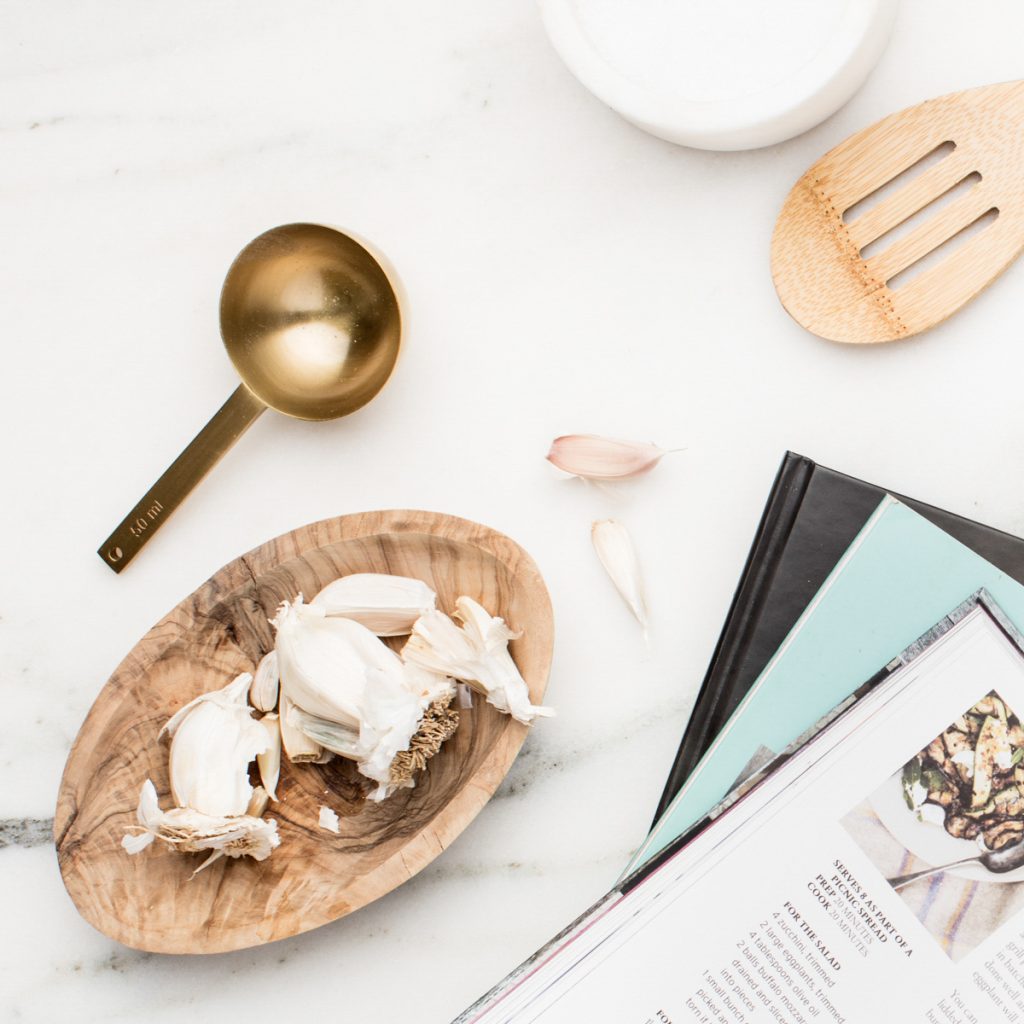 A flat lay of garlic cloves on a wooden dish, a gold measuring cup, a wooden slotted spoon, and cookbooks on a white marble surface. Parts of a recipe page are also visible.