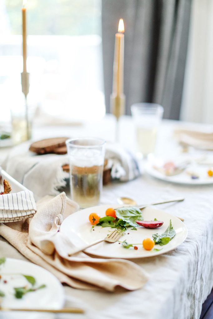 A dining table set with white plates, gold cutlery, and beige napkins. Plates hold salad greens and cherry tomatoes. Lit candles and a glass with iced drink are also on the table, creating a cozy, elegant atmosphere.