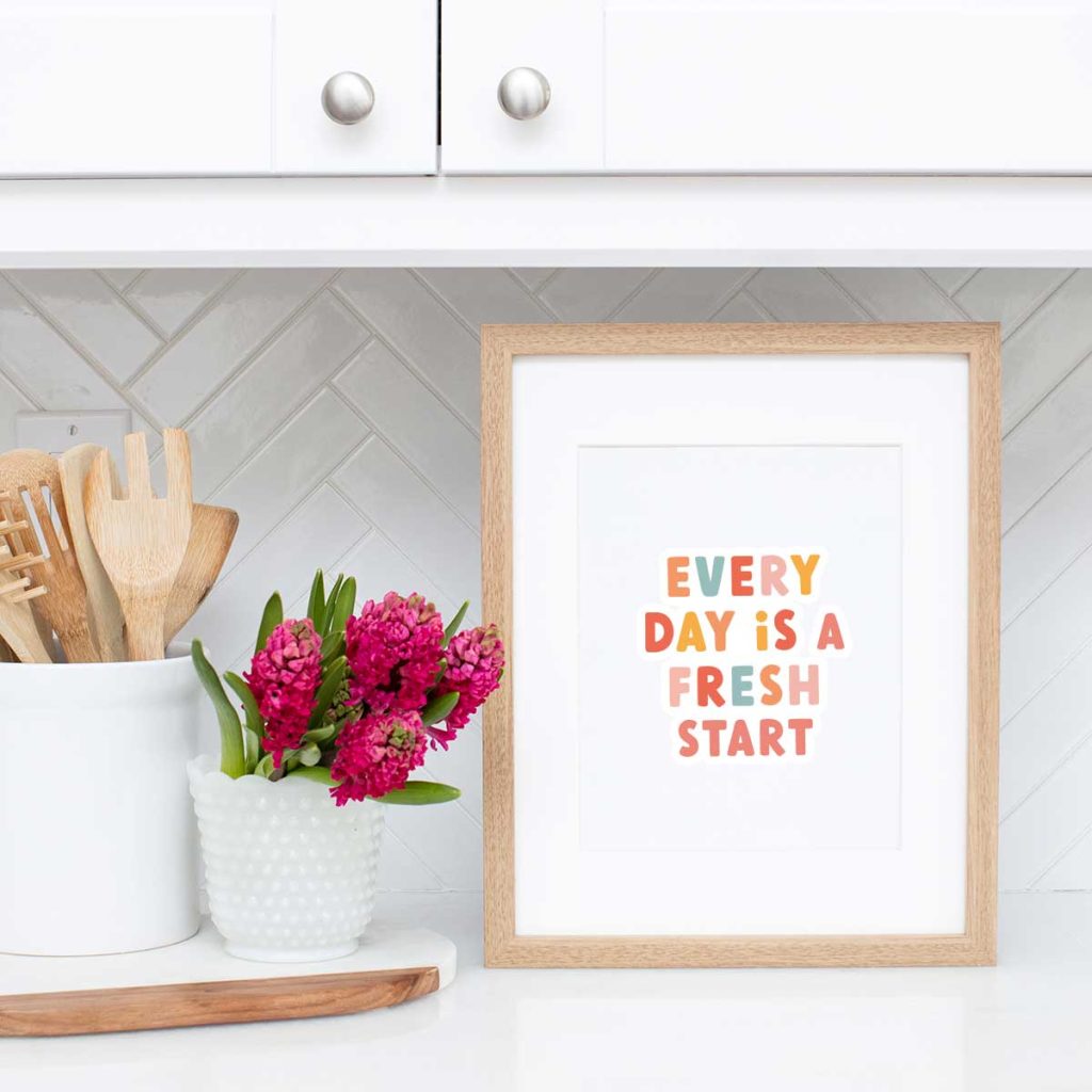 A framed print with the words “EVERY DAY IS A FRESH START” sits on a white kitchen counter next to a white vase of pink flowers and a white container holding wooden utensils.