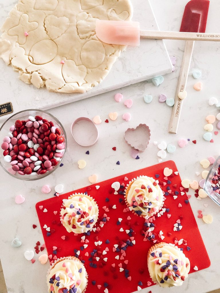 A baking scene with cupcakes topped with heart-shaped sprinkles, cookie dough being cut into heart shapes, bowls of red candies, and a spatula on a white countertop decorated with colorful heart sprinkles.