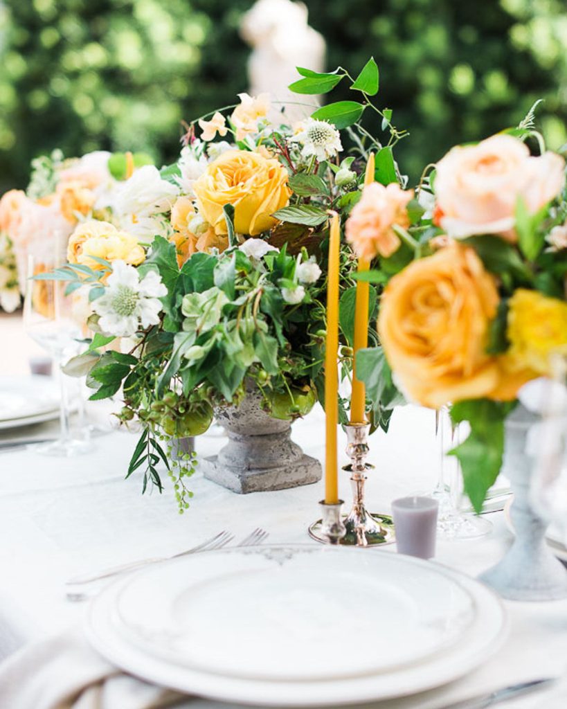 A beautifully set outdoor table features elegant white plates, silver cutlery, yellow and peach rose floral centerpieces, lush greenery, and tall yellow candles in silver holders. The background is softly blurred with greenery.