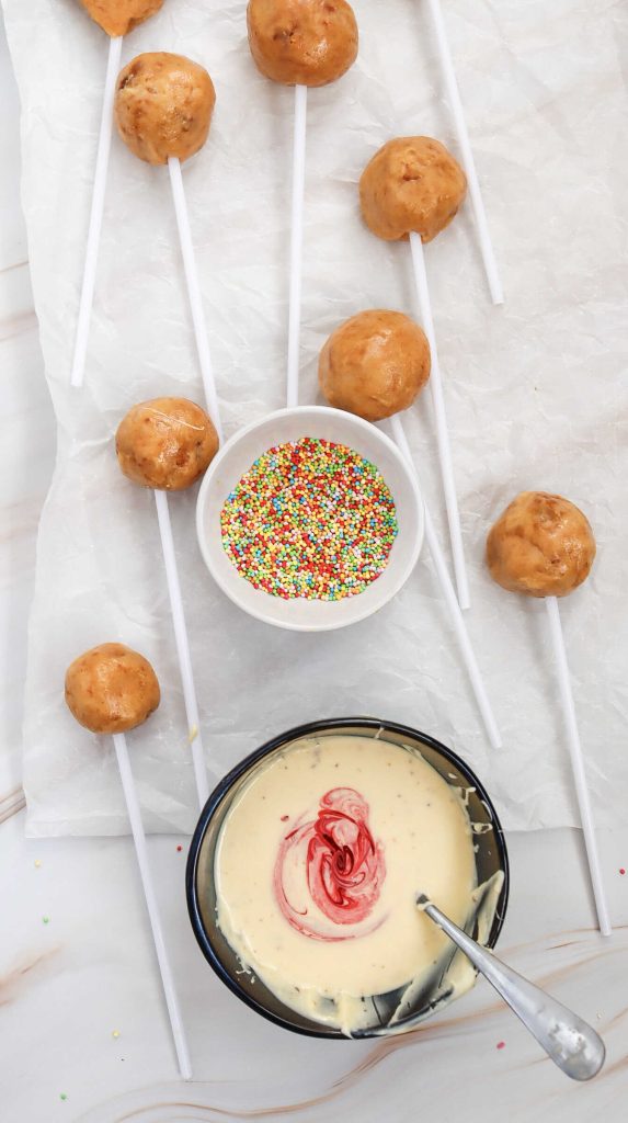 Easy cake pops on sticks are laid out on parchment paper. A small bowl of colorful sprinkles and a larger bowl of white melted chocolate with red coloring swirled in are nearby, with a spoon in the chocolate.