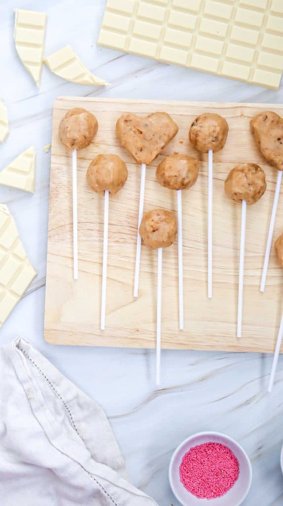 A wooden board with round and heart-shaped easy cake pops on sticks, surrounded by white chocolate pieces, a small bowl of pink sprinkles, and a white cloth on a marble surface.