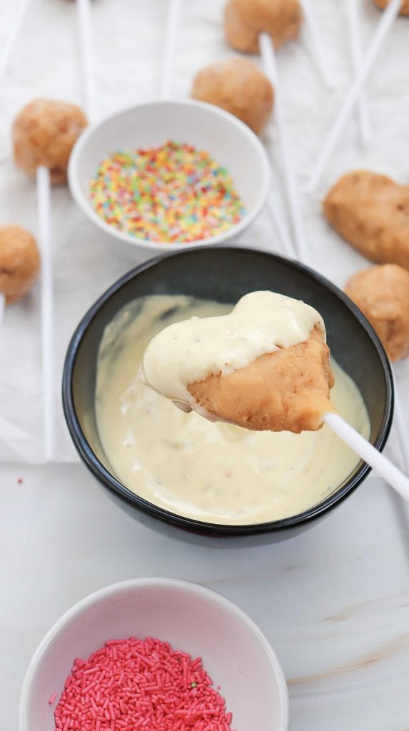 A cake pop on a stick being dipped into white chocolate, with bowls of colorful sprinkles and more easy cake pops on sticks in the background.