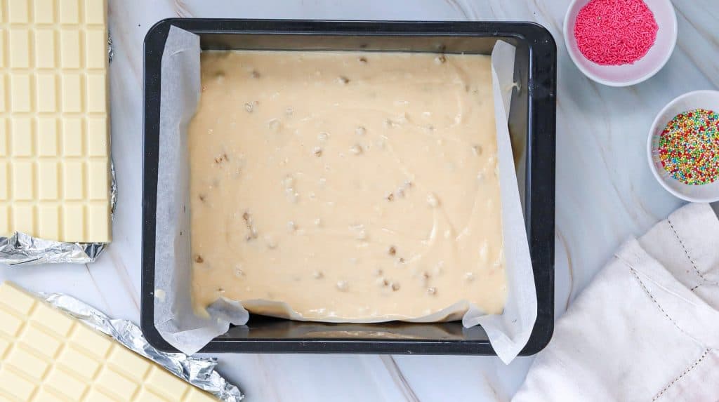 A square baking pan lined with parchment paper holds unbaked cookie dough on a marble surface. Nearby are blocks of white chocolate, a white napkin, and bowls of pink and rainbow sprinkles—perfect for making easy cake pops.