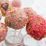 Easy cake pops covered in pink, red, and white icing are topped with multicolored sprinkles and arranged on white sticks in a glass jar. A blurred red heart decoration adds a festive touch in the background.
