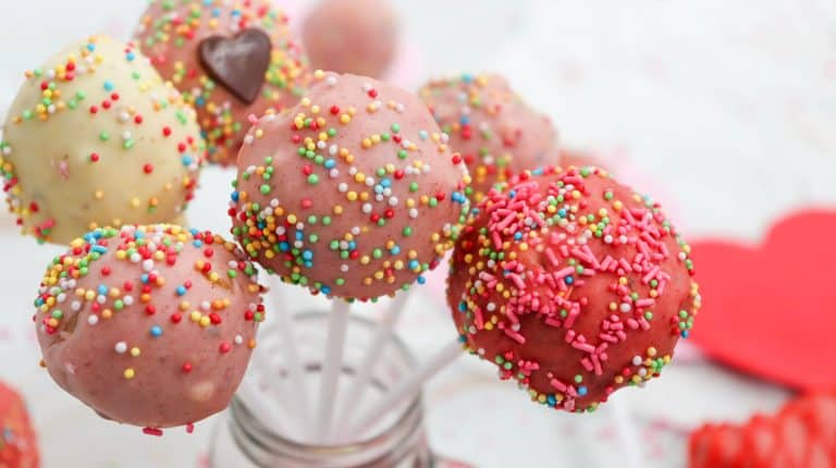 Easy cake pops covered in pink, red, and white icing are topped with multicolored sprinkles and arranged on white sticks in a glass jar. A blurred red heart decoration adds a festive touch in the background.