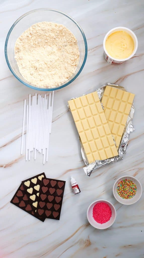 A flat lay of baking ingredients and supplies for easy cake pops on a marble surface, including cake mix in a bowl, tub of yellow frosting, white chocolate bars, heart-shaped chocolate, cake pop sticks, food coloring, and bowls of sprinkles.