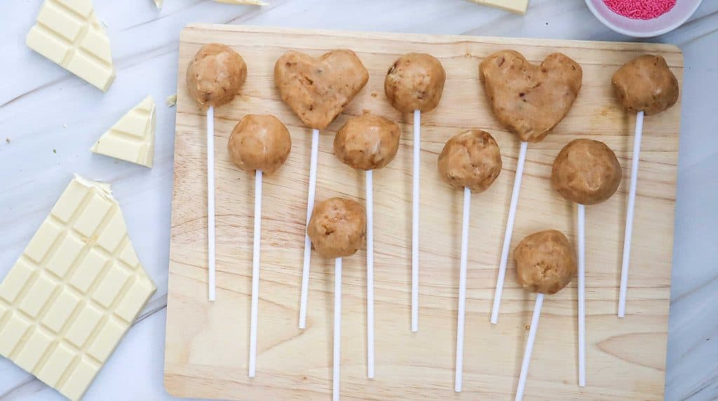 A wooden board displays easy cake pops made from round and heart-shaped cookie dough on sticks. Nearby, white chocolate bars and a bowl of pink sprinkles rest on a white marble surface.