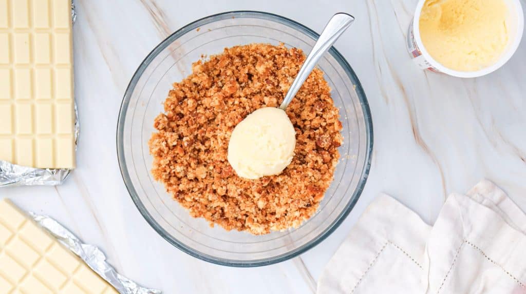 A bowl of crumbly brown mixture with a scoop of vanilla ice cream on top and a spoon, next to a container of ice cream, a white chocolate bar, easy cake pops, and a white napkin on a marble surface.