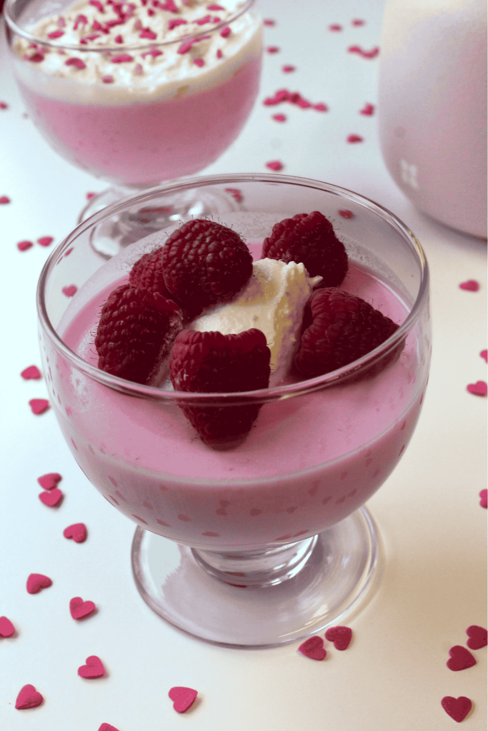A glass dessert cup filled with pink mousse, topped with whipped cream and fresh raspberries—one of the best Valentine's Day desserts. Small pink heart-shaped sprinkles decorate the white surface, with another dessert cup blurred in the background.