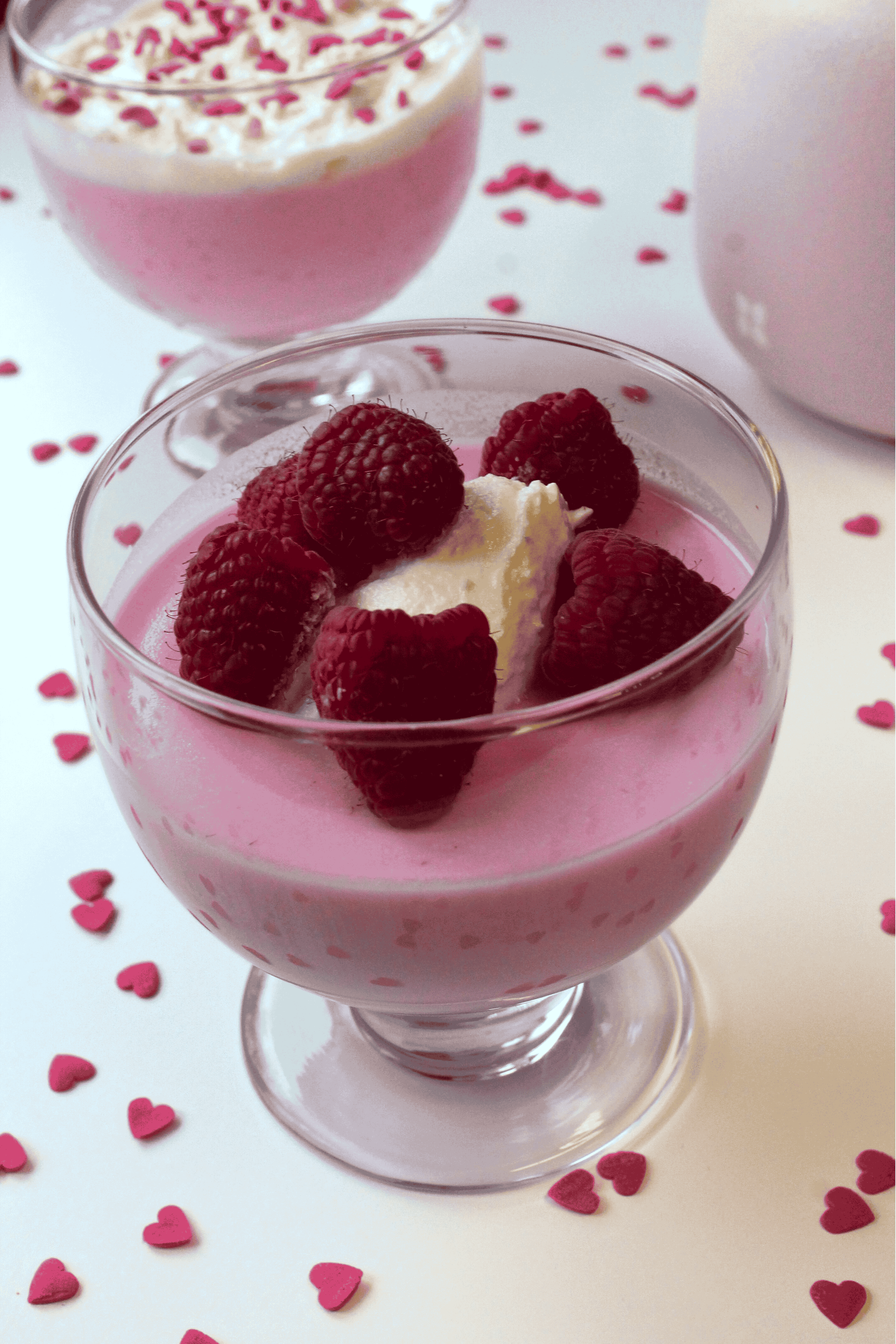 A glass dessert cup filled with pink mousse, topped with whipped cream and fresh raspberries—one of the best Valentine's Day desserts. Small pink heart-shaped sprinkles decorate the white surface, with another dessert cup blurred in the background.