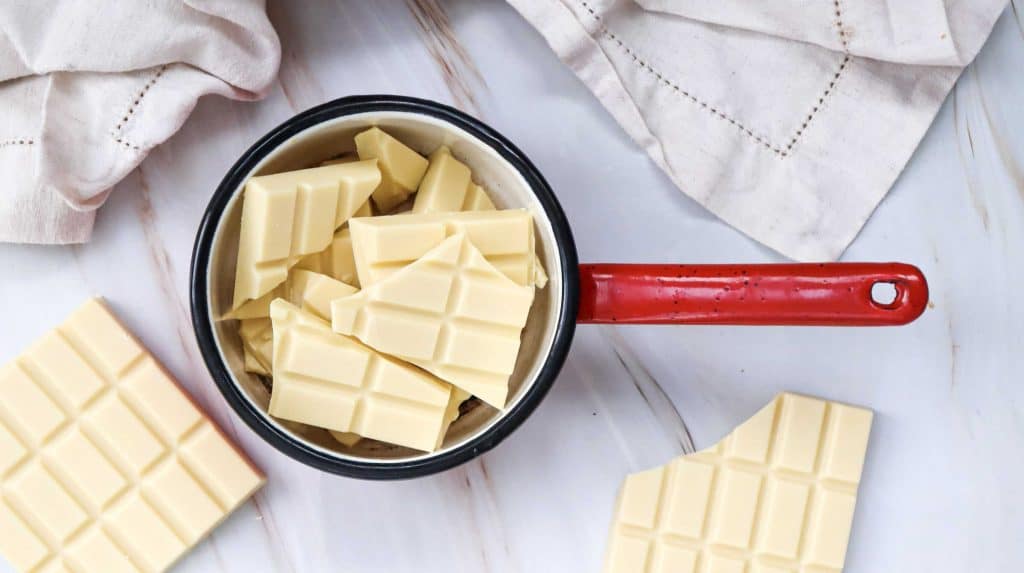 A saucepan with a red handle filled with broken pieces of white chocolate sits on a white surface, surrounded by cream-colored cloths and more white chocolate pieces—perfect for melting chocolate for easy cake pops.