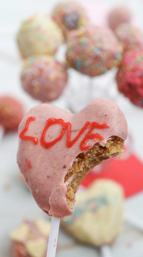 A heart-shaped easy cake pop coated in pink icing with the word “LOVE” written in red, partially bitten and held on a stick. Blurry, colorful cake pops are visible in the background.
