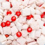 A clear bowl filled with white, powdered cereal squares and scattered red M&M candies, this strawberry puppy chow snack mix is the perfect sweet treat for any occasion.