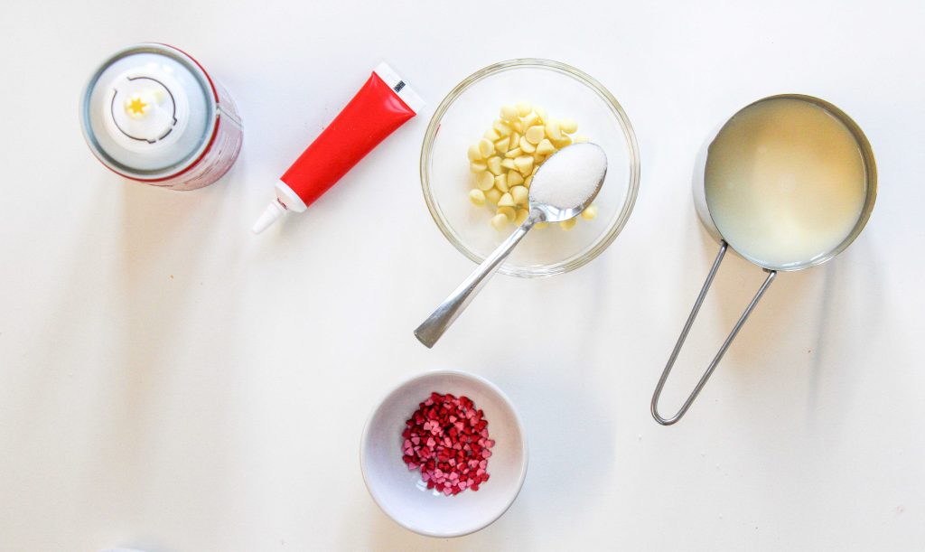 Ingredients for a red velvet hot chocolate drink on a white surface: whipped cream can, red icing tube, bowl of white chocolate chips with a spoonful of sugar, metal measuring cup with milk, and a small bowl of red sprinkles.