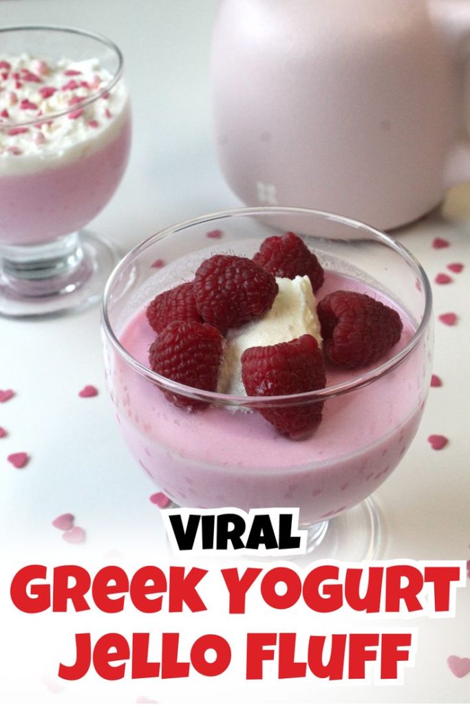 A glass bowl filled with pink Greek Yogurt Jello Fluff, topped with whipped cream and fresh raspberries. Another bowl and a pink pitcher are blurred in the background. Heart-shaped sprinkles are scattered on the white surface.