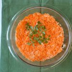 A glass bowl filled with grated carrots and onions, garnished with fresh parsley, sits on a green cloth napkin—an easy carrot recipe perfect as a carrot side dish.