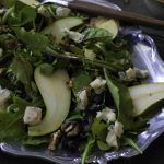 A pear walnut blue cheese salad with mixed greens is served on a silver platter, garnished with crumbled blue cheese and walnuts, a wooden spoon beside it, and a bowl of dressing nearby on a dark surface.