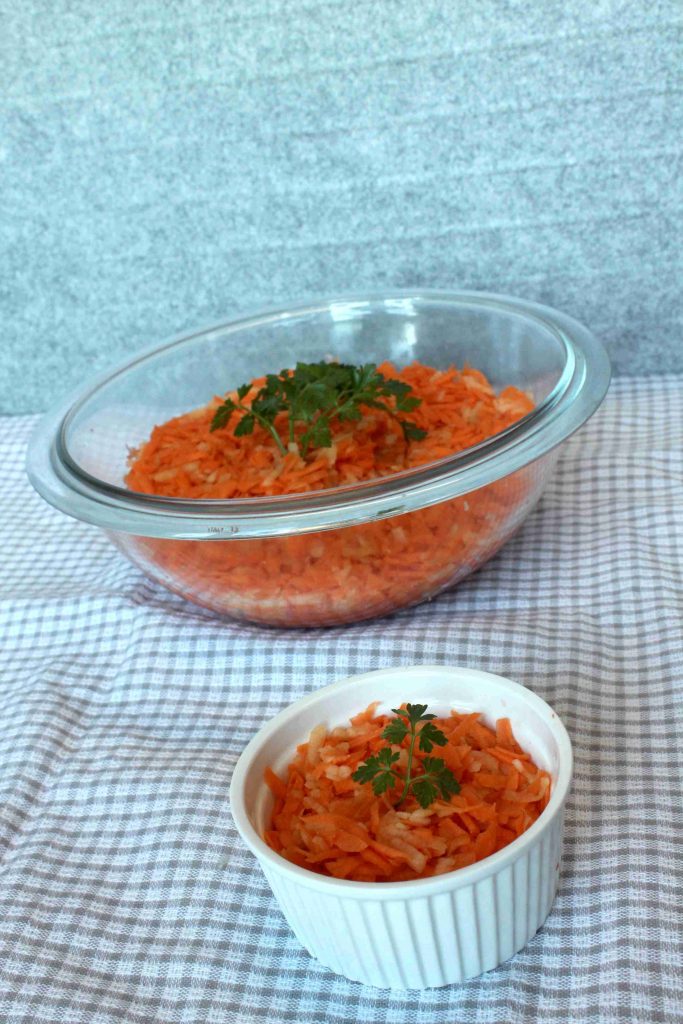 A large glass bowl and a small white ramekin filled with grated carrots, each garnished with parsley, sit on a checkered cloth against a light gray background — a perfect kid friendly carrots side dish for any meal.