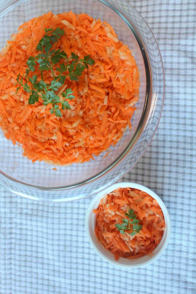 An easy carrot side dish, perfect for kids, is served in a glass bowl and a small white bowl, both filled with grated carrot and onion salad garnished with fresh parsley on a checkered tablecloth.