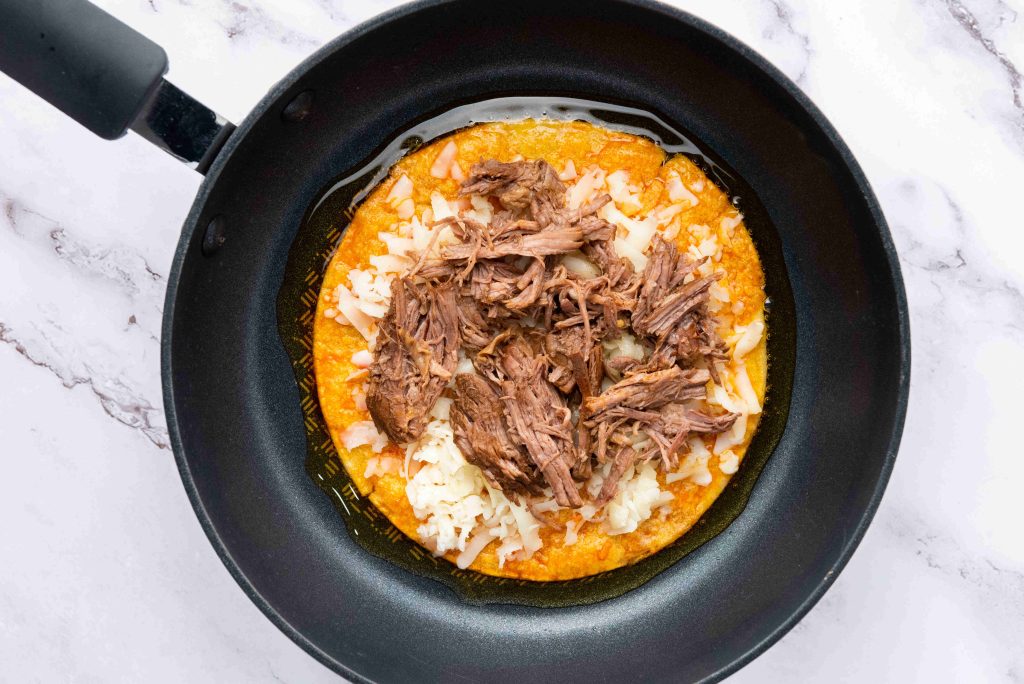 A corn tortilla topped with melted cheese and shredded beef, inspired by a birria tacos recipe, sits in a black skillet on a white marble surface.