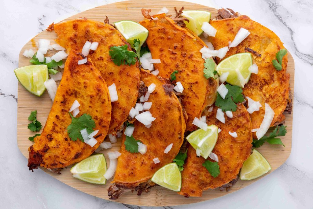 Five crispy birria tacos, made using our savory birria tacos recipe, are filled with shredded meat, topped with onions and cilantro, and served on a wooden platter with lime wedges on a marble surface.