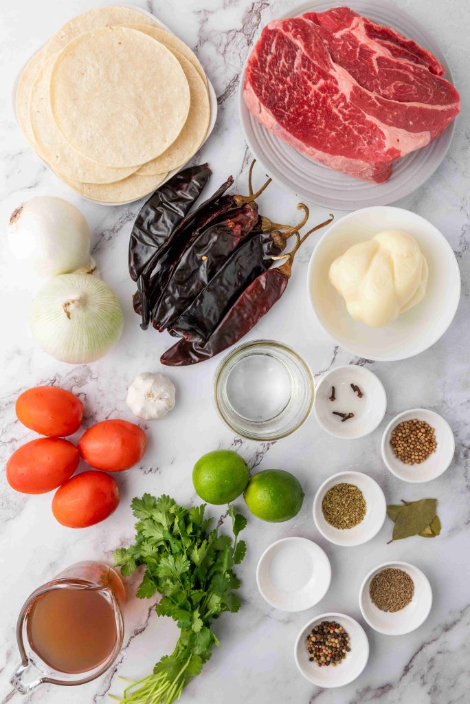 Overhead view of ingredients on a marble surface for a birria tacos recipe: raw beef, corn tortillas, dried chiles, white onions, tomatoes, garlic, limes, fresh cilantro, bay leaves, whole spices, salt, pepper, vinegar, and broth.