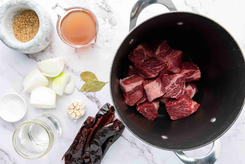 Chunks of raw beef in a black pot surrounded by onion, dried chili peppers, garlic, bay leaves, spices, broth, and water on white marble—perfect ingredients for an authentic birria tacos recipe.