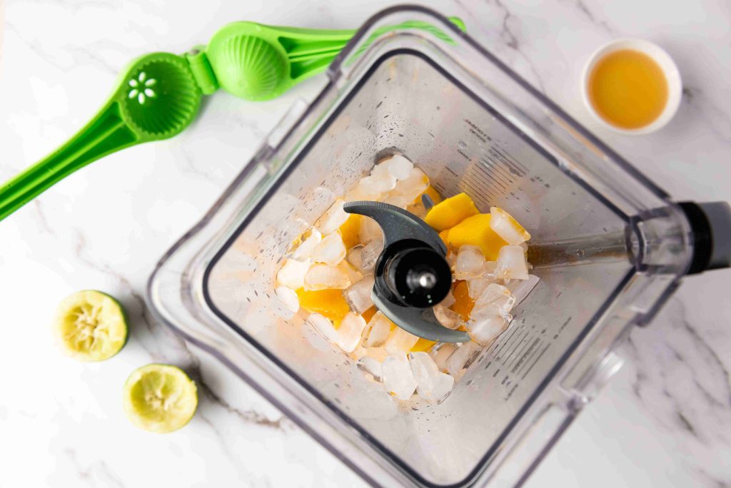 Top-down view of a blender filled with ice cubes and mango pieces, ready to make a Frozen Mango Margarita. Nearby are a green citrus squeezer, two squeezed lime halves, and a small bowl of juice on a white marble surface.