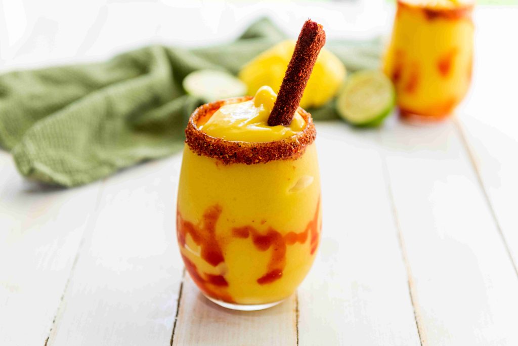 A glass of bright yellow mangonada with red sauce swirls, rimmed with chili powder, and garnished with a tamarind candy stick. This sweet and spicy drink is an easy mangonada to enjoy anytime. Another glass and green cloth are blurred in the background.