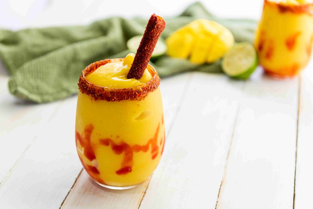 A glass of mangonada topped with a tamarind stick and chili powder rim, swirled with chamoy sauce—an easy mangonada that’s the perfect sweet and spicy drink. Chopped mango, lime, and a green cloth sit on a white wooden surface in the background.