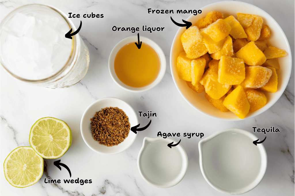 Overhead view of ingredients for a Frozen Mango Margarita in bowls and glasses on a marble surface, labeled: ice cubes, frozen mango, orange liquor, Tajin, agave syrup, tequila, and lime wedges.