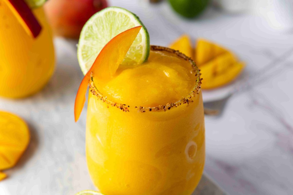 A yellow Frozen Mango Margarita in a glass with a chili powder rim, garnished with a lime wheel and mango slice. Slices of mango and lime are nearby on a white surface.