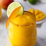 A yellow Frozen Mango Margarita in a glass with a chili powder rim, garnished with a lime wheel and mango slice. Slices of mango and lime are nearby on a white surface.