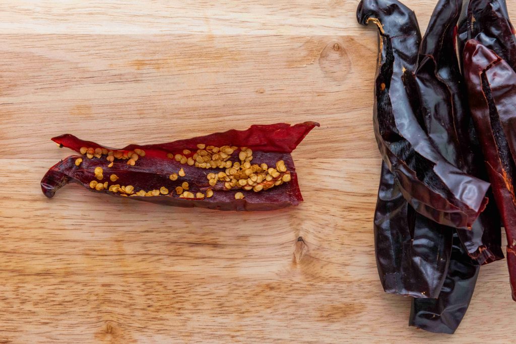 A dried red chili pepper split open with seeds visible on a wooden surface, next to a small pile of whole dried chili peppers—essential ingredients for an authentic birria tacos recipe.
