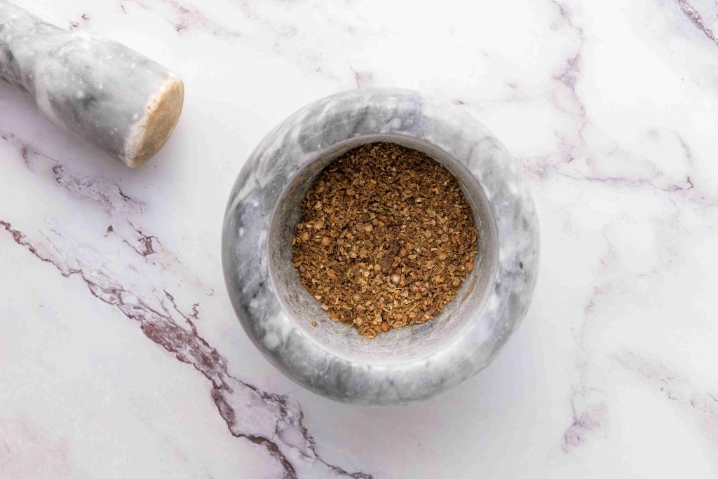 A marble mortar and pestle on a white marble surface, with the mortar containing ground brown spices—perfect for preparing spices for your favorite birria tacos recipe.