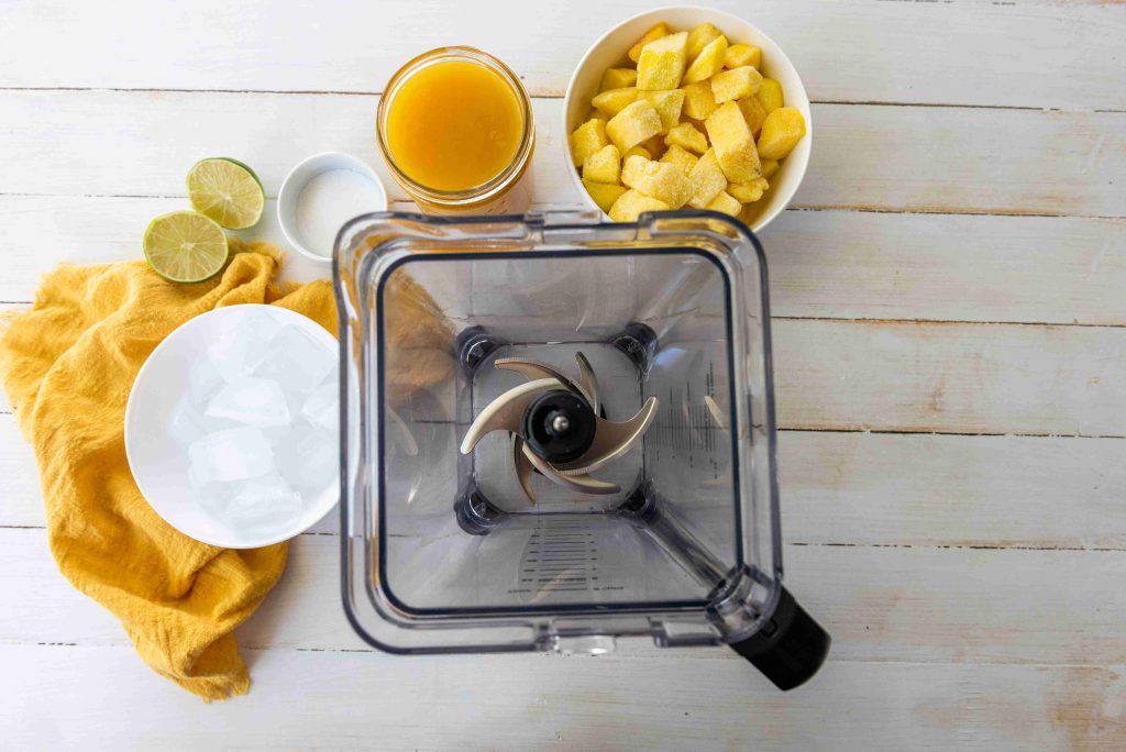 A top view of a blender surrounded by a bowl of ice, lime halves, a jar of orange juice, and chopped mango on a white wooden surface with a yellow cloth—perfect for an easy mangonada or sweet and spicy drink.