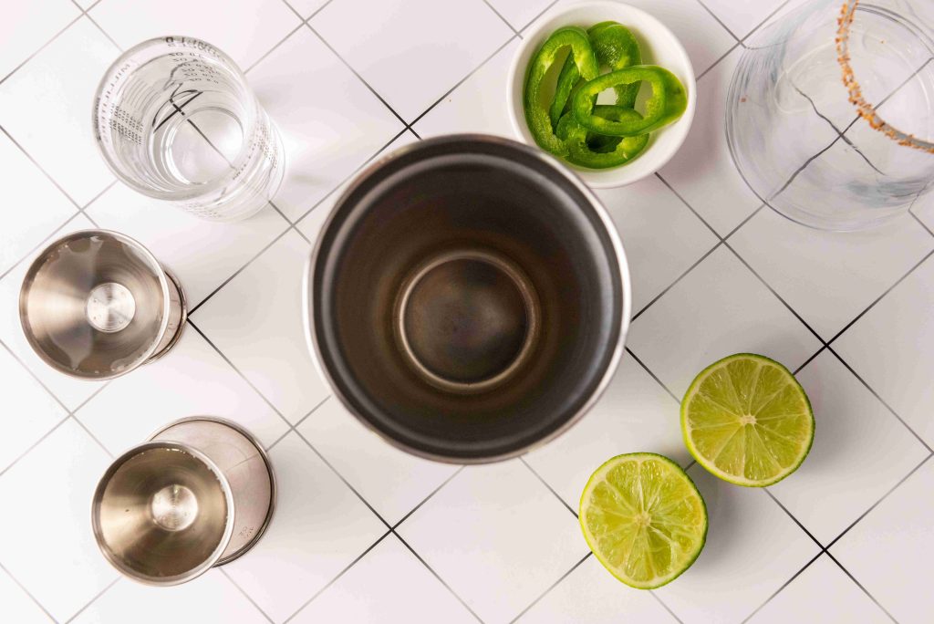A top-down view of a cocktail shaker, jiggers, a glass, a bowl of sliced green peppers, and two lime halves on a white tiled surface—perfect essentials for crafting a Spicy Margarita or Jalapeno Tequila cocktail.